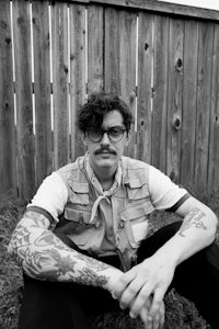 a man with tattoos sitting in front of a fence